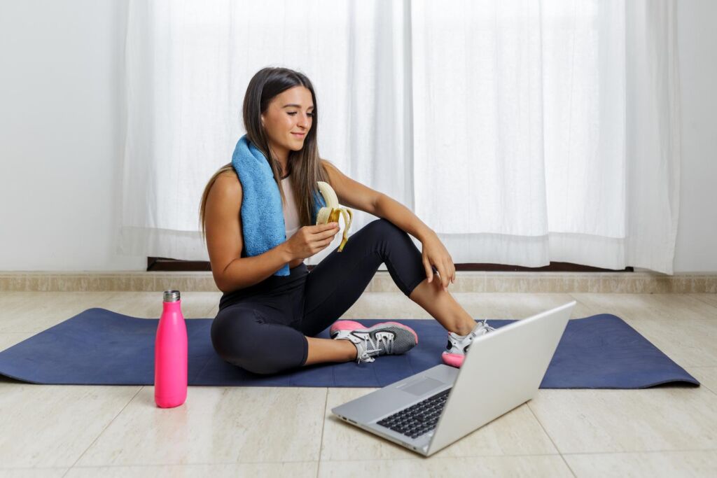 sportswoman sitting on mat and eating banana after online training